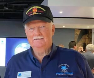 Portrait of Fred Huskey, an elderly man with white hair and a mustache, smiling gently while seated indoors, wearing a brown polo shirt.
