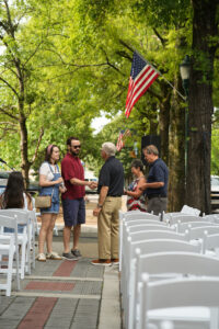 People greet each other in a city plaza surrounded by trees and an American flag in waving in the distance.
